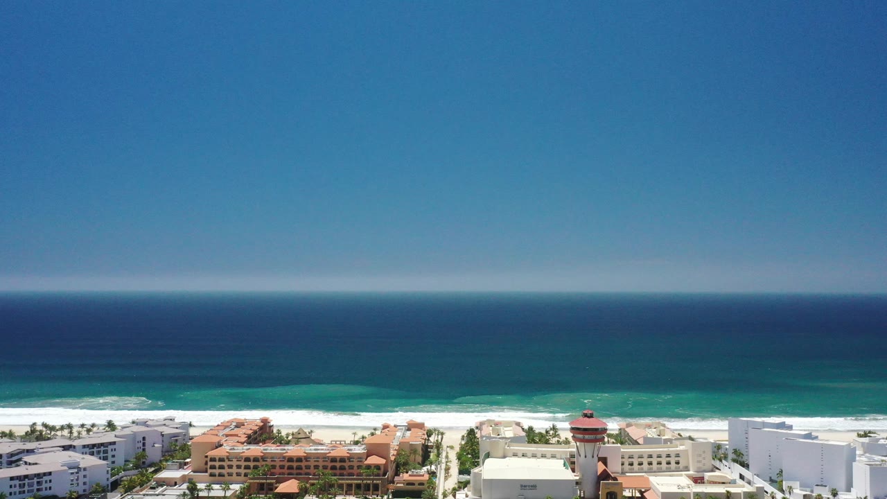 San Jose Del Cabo coastline from above, with layered blue water, a white surf line, pale sand, and resort roofs slightly more centered at the bottom, with no visible people.