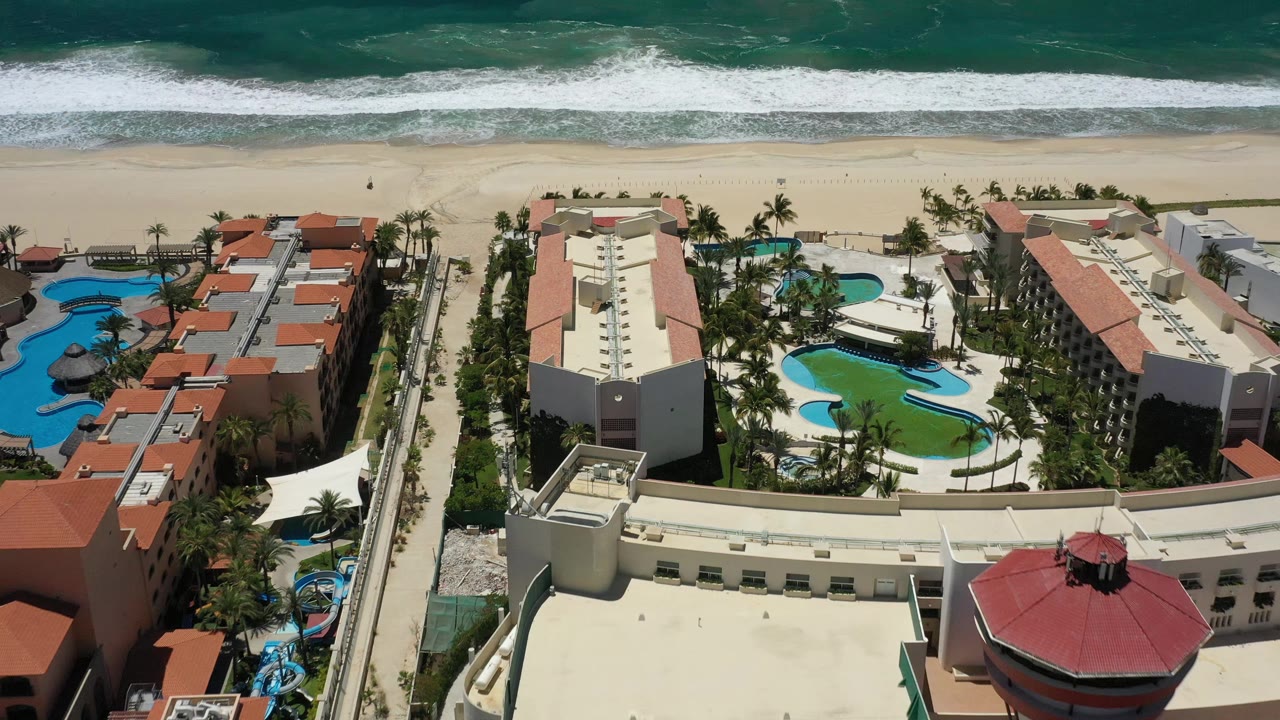 Aerial view in San Jose del Cabo showing resort rooftops and pool courtyards below a wide sand beach and a horizontal band of white breaking waves.