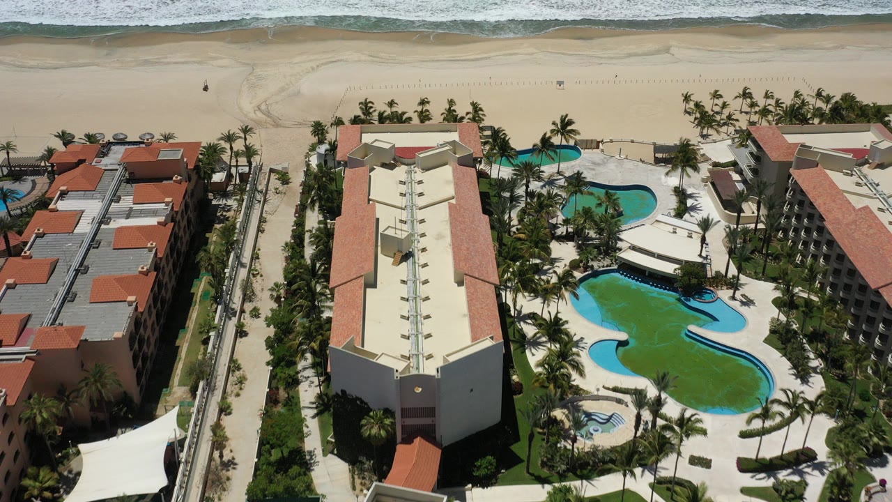 Overhead drone view of a San Jose del Cabo beachfront hotel with red roofs, curving pools, palm landscaping, sandy beach, and no visible people.