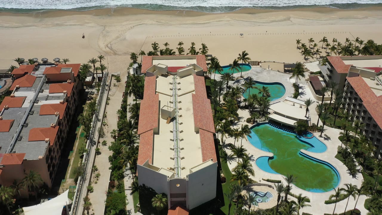 Aerial hotel courtyard with pools, palms, adjacent buildings, beach, and waves.