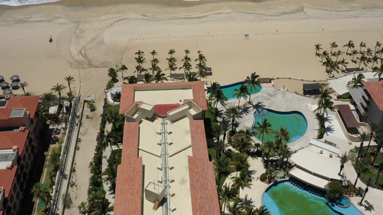 High aerial view in San Jose del Cabo showing red tile resort roofs at the bottom, turquoise pools among palms, broad beach sand, and surf along the top edge.