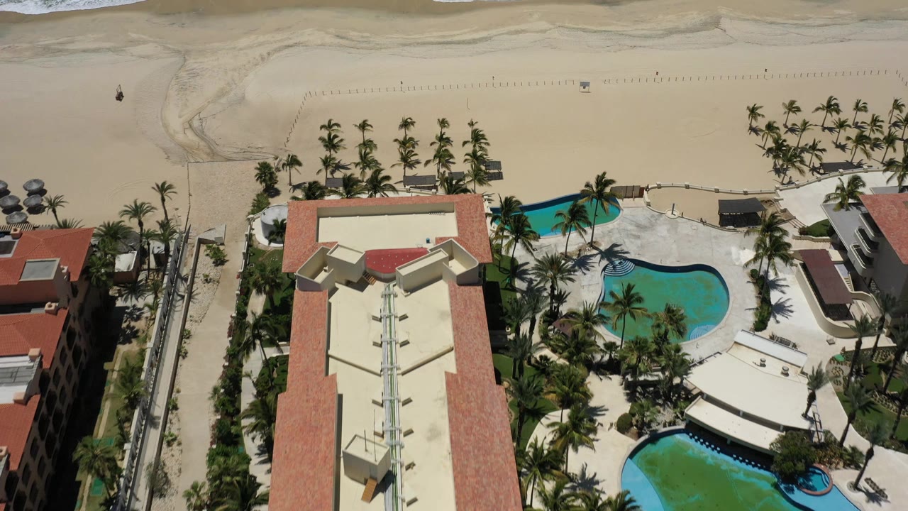 Drone-height frame in San Jose del Cabo with the central resort roof, curved blue pools, rows of palms, pale sand, and a small surf line beyond the beach.