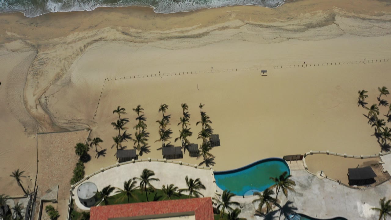 Overhead view in San Jose del Cabo, Mexico, showing beachhouse terrace edges, a curved blue pool, palm clusters, pale sand, and surf at the top with no visible people.