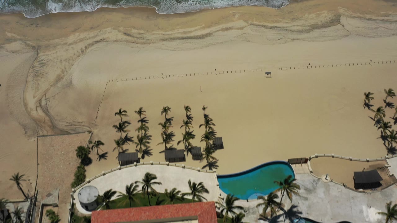 Aerial top-down frame in San Jose del Cabo, Mexico, with the pool and palm-lined terrace still visible below a broad empty sand area and foamy shoreline, with no visible people.