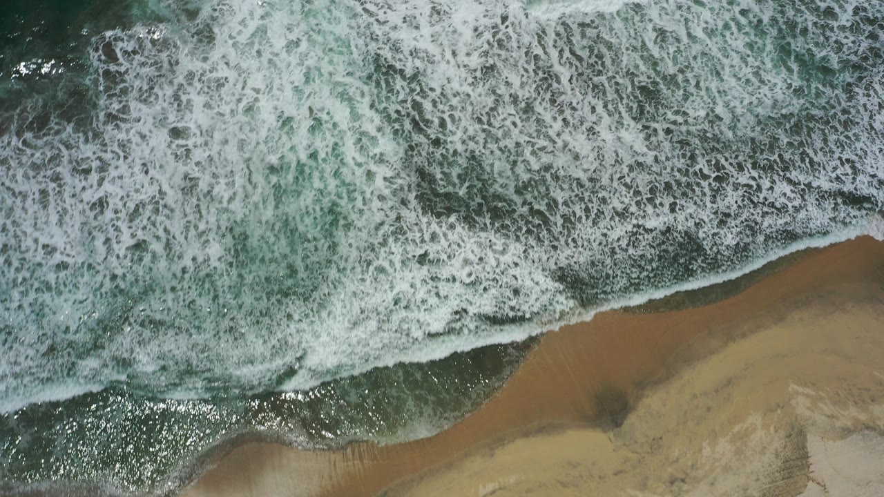 Overhead view of white surf meeting tan beach in San Jose Del Cabo, Mexico, with dark green water and no visible people.