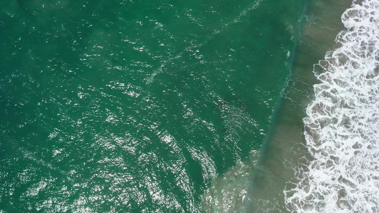 Aerial frame of San Jose Del Cabo ocean water with shimmering green-blue surface, a foamy wave band at right, and no visible people.