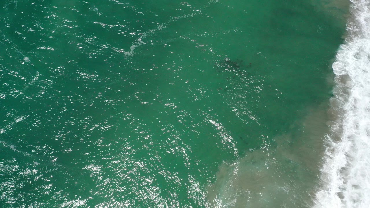 Overhead view of teal ocean water in San Jose del Cabo, Mexico, with sun glints across the surface, pale shallows near the surf, and no visible people.