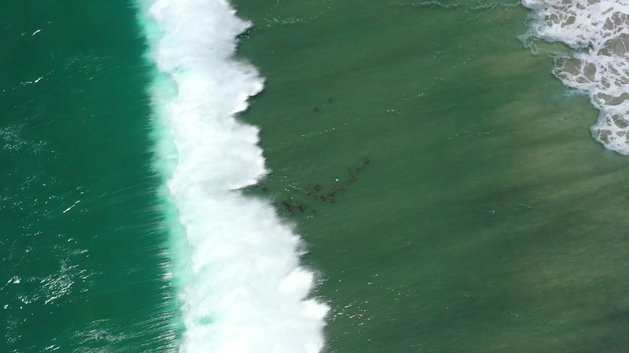 Overhead view of green ocean water and a narrow white surf line over sandy shallows in San Jose del Cabo, Mexico, with no visible people.