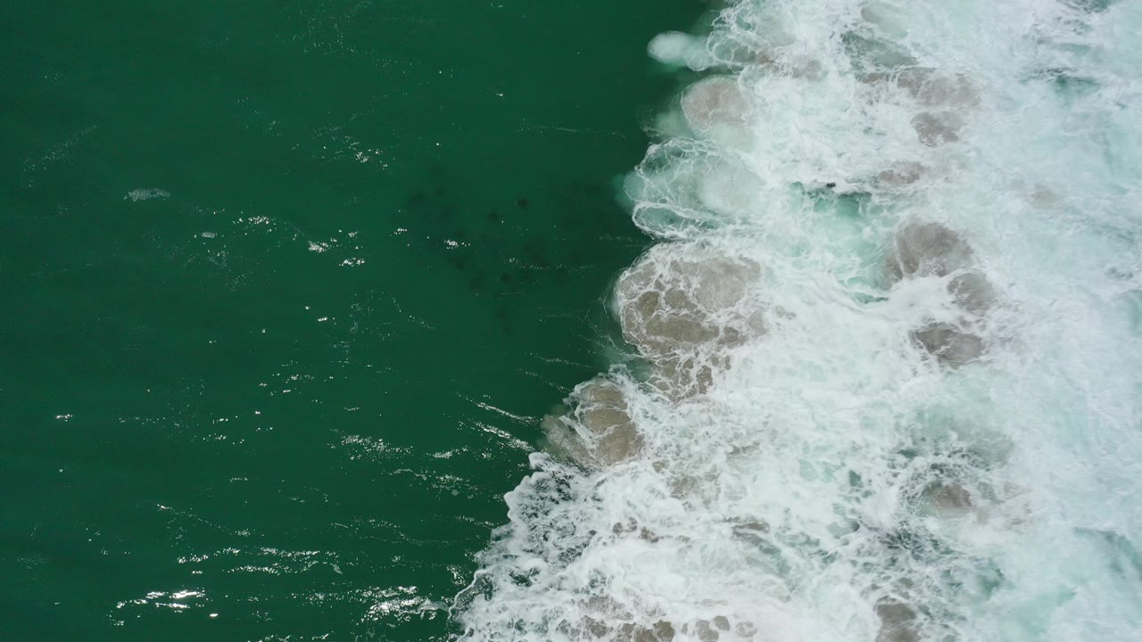 At 0:00, an overhead view in San Jose del Cabo shows white surf entering from the right over green-turquoise water with dark submerged fish shapes and no visible people.