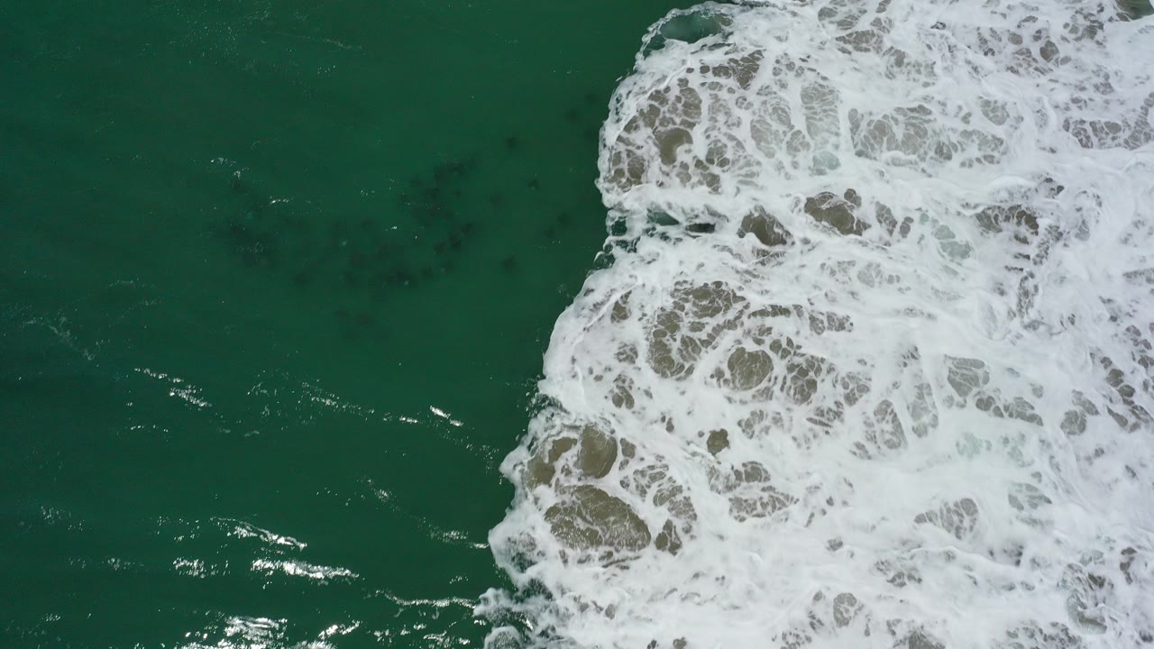 Aerial view over green ocean water in San Jose Del Cabo, Mexico, with white surf foam entering from the right side and no visible people.