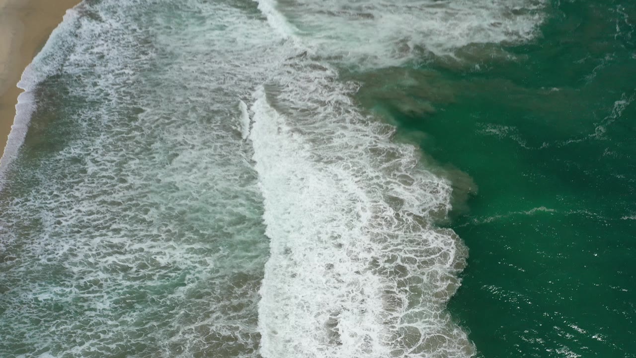 Tan sand appears along the left edge while white surf spreads through green water in San Jose del Cabo, with no visible people on the beach or in the water.