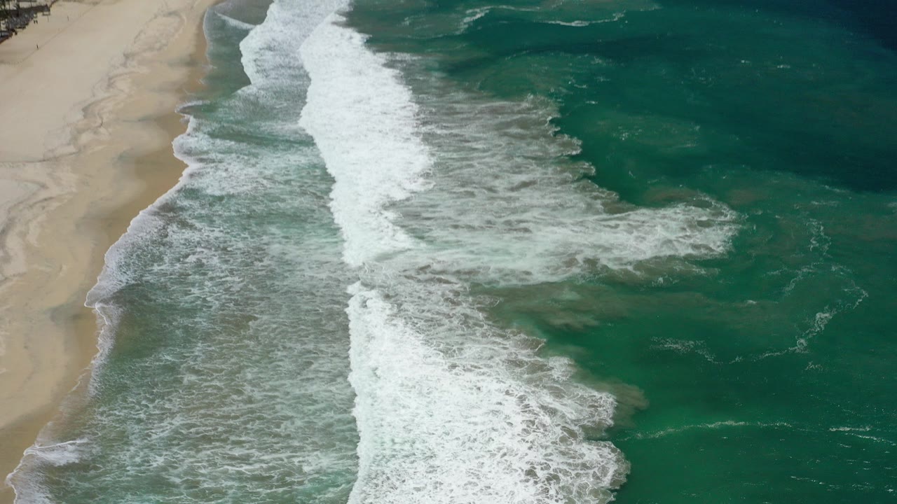Aerial view of surf breaking along a pale sand beach.