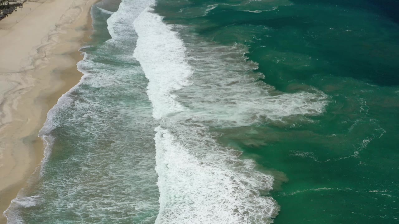Aerial view over San Jose del Cabo beach with a stronger diagonal surf line, textured shallow water near shore, darker green water offshore, and no visible people.