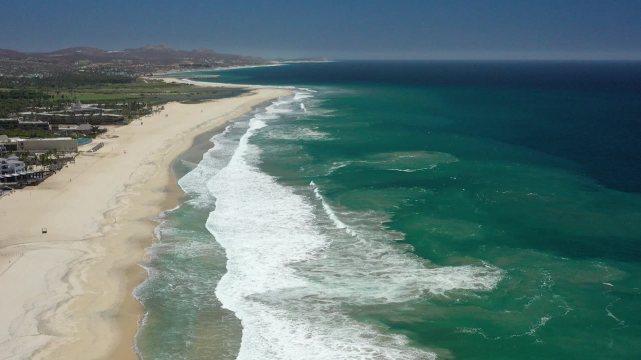 Opening aerial view of San Jose del Cabo coastline with a wide sandy beach on the left, white surf spreading into turquoise water, coastal buildings near the lower left, distant hills, and no visible people.