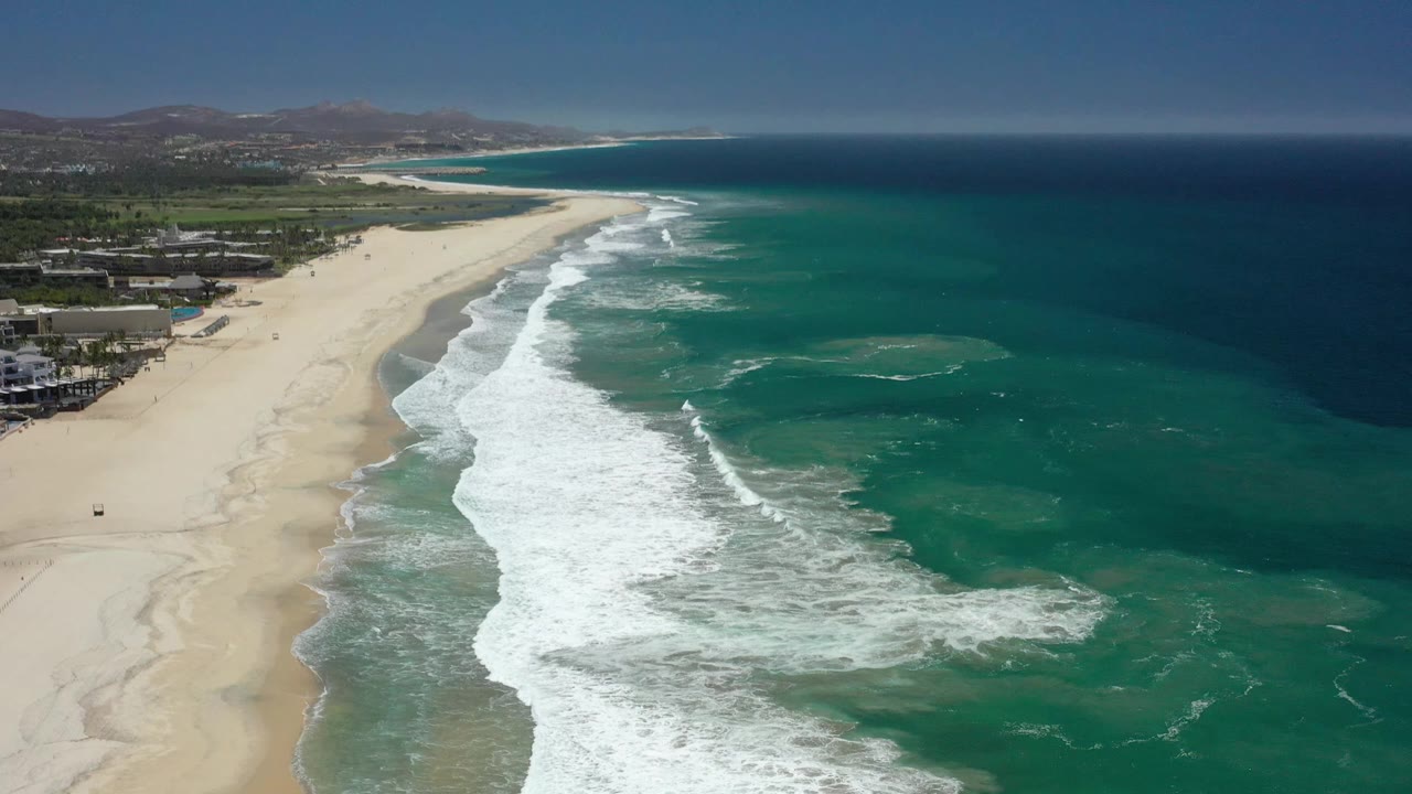 Aerial frame over San Jose del Cabo showing pale sand, a long white surf break, turquoise nearshore water, darker ocean beyond, low buildings at the beach edge, hills in the background, and no visible people.