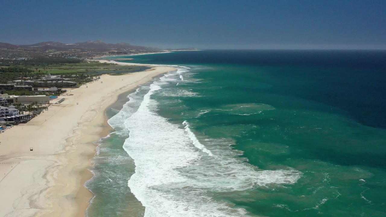 Diagonal aerial view along a long sandy coast with foamy waves and open ocean.