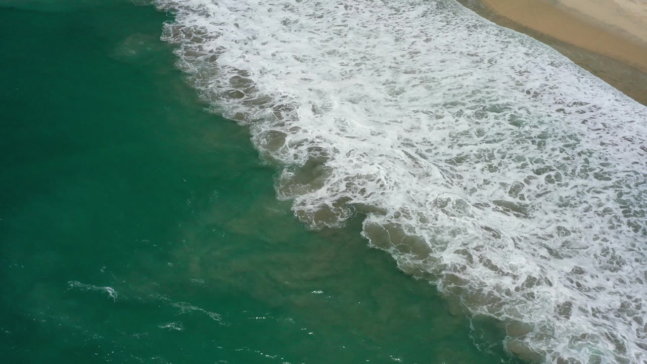 Top-down view in San Jose Del Cabo, Mexico, showing deep green water and white surf curling diagonally toward tan beach, with no visible people.