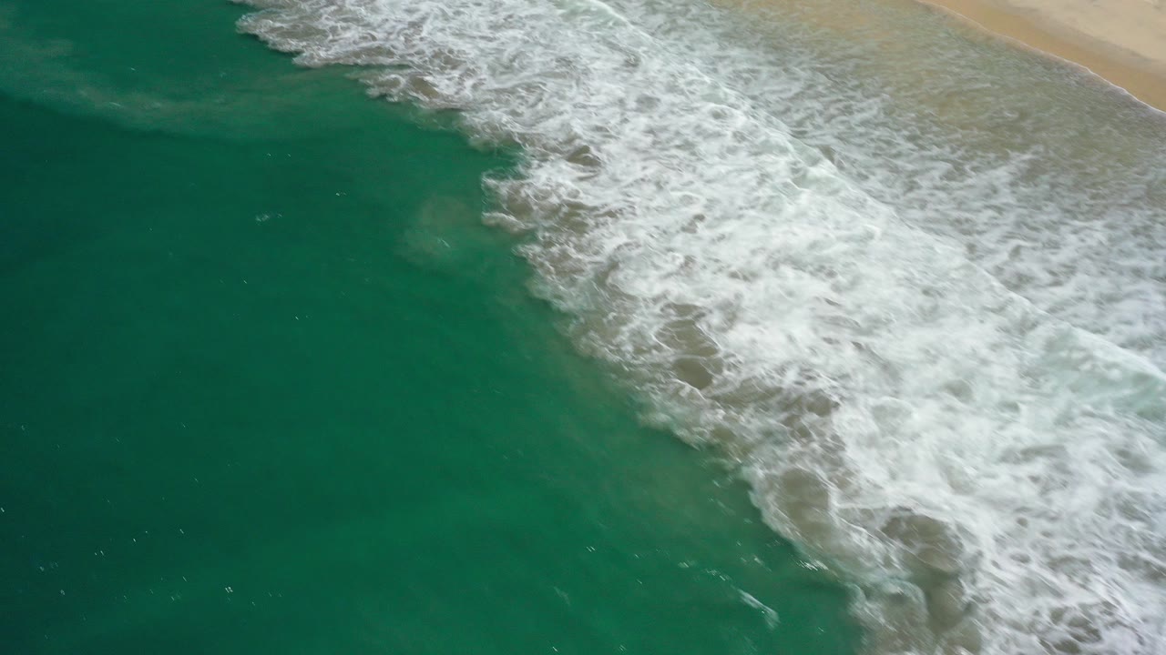 Aerial view over San Jose Del Cabo ocean water as white surf spreads diagonally toward a pale sandy beach, with no visible people.