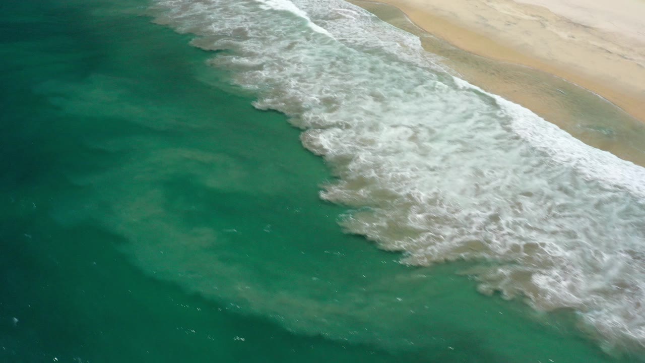 Aerial view in San Jose Del Cabo, Mexico showing green-blue water and a broad band of white surf reaching pale beach sand, with no visible people.