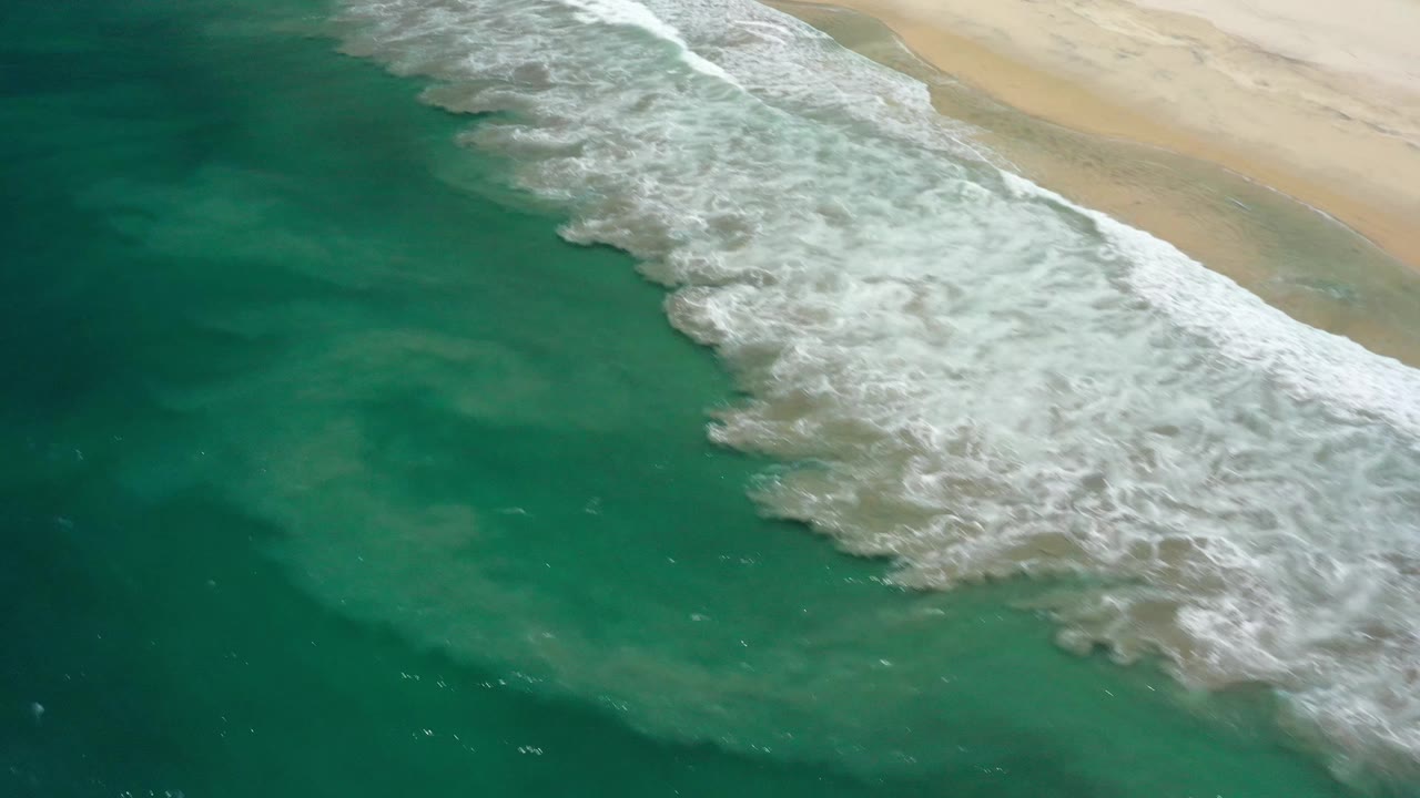 Aerial shoreline frame in San Jose Del Cabo, Mexico with foamy white water spreading over green sea beside pale sand, with no visible people.