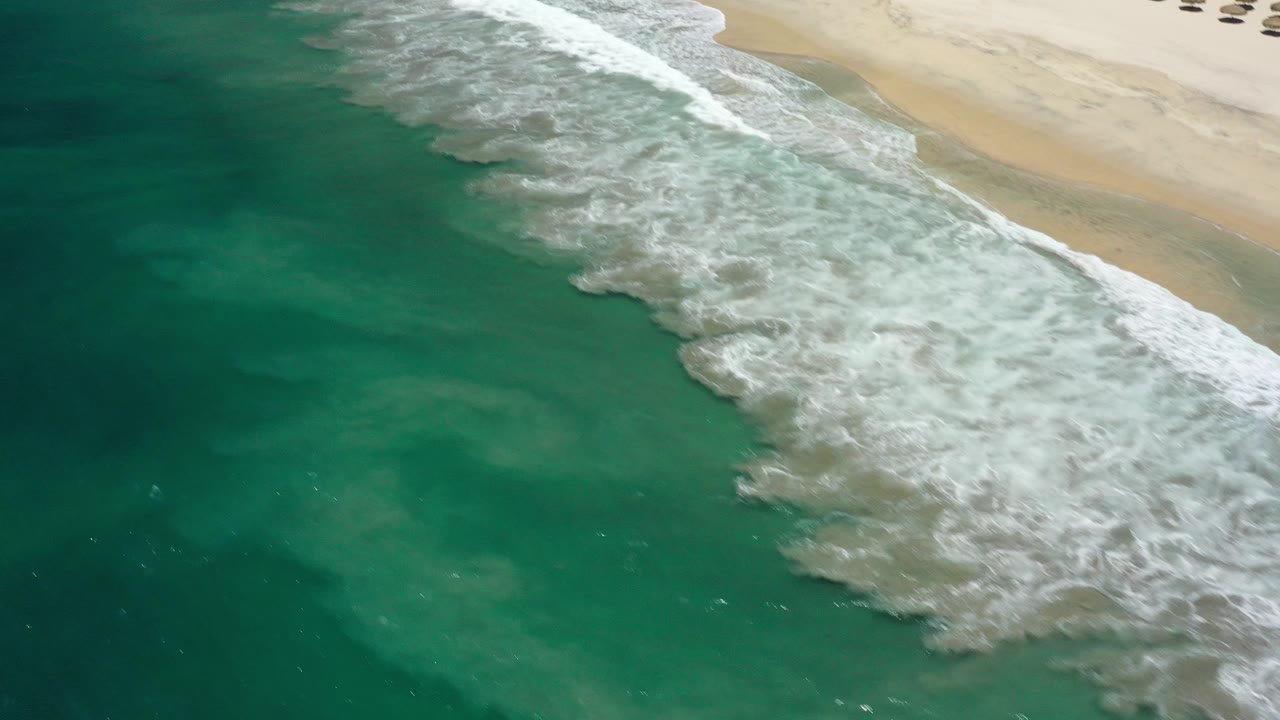 Aerial view in San Jose Del Cabo, Mexico with turquoise water, a diagonal break of white surf, pale sand, and small beach umbrellas near the upper edge, with no visible people.