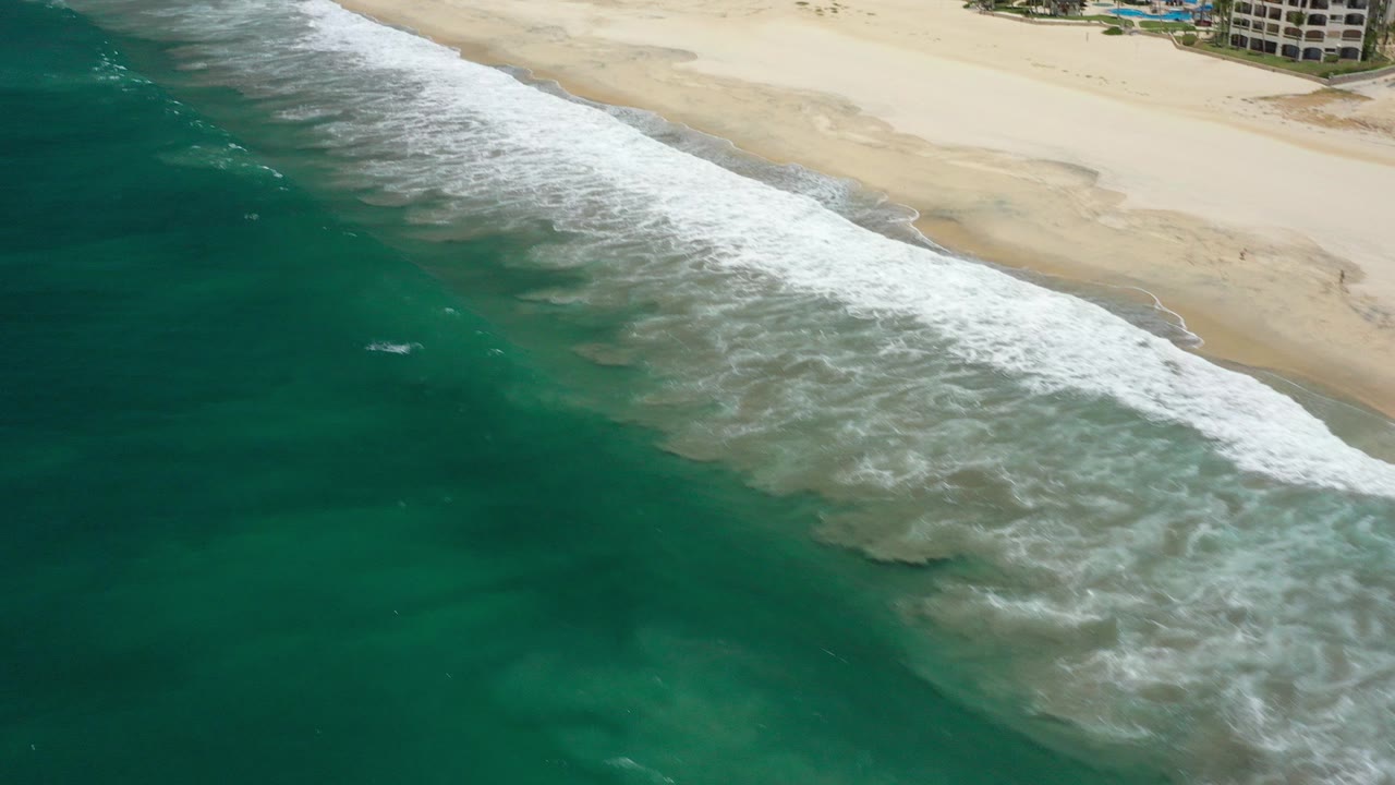 Aerial view over San Jose del Cabo where turquoise water and white surf meet a wide sandy beach, with no visible people in the frame.