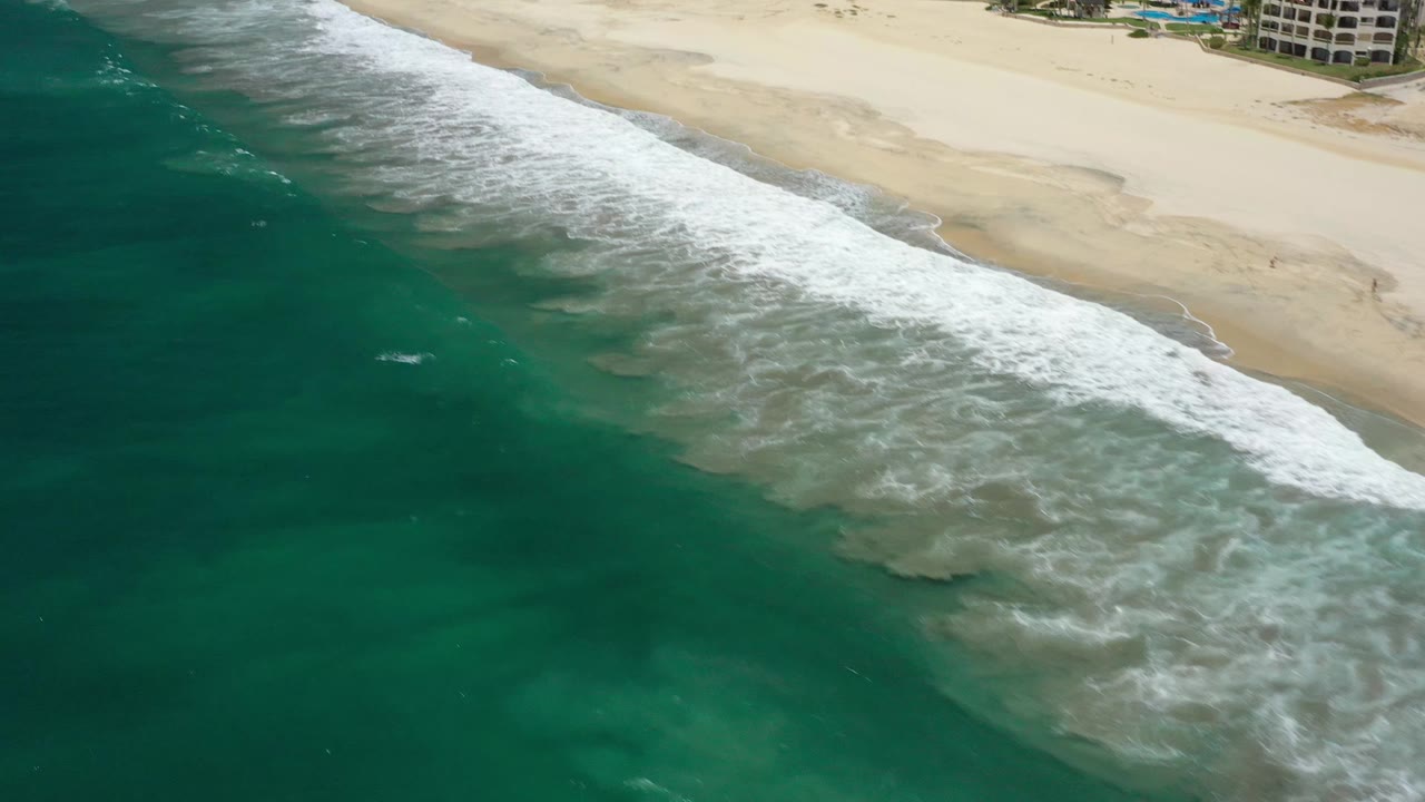 White foam spreads across shallow turquoise water beside the pale San Jose del Cabo beach, with distant buildings at the top edge and no visible people.