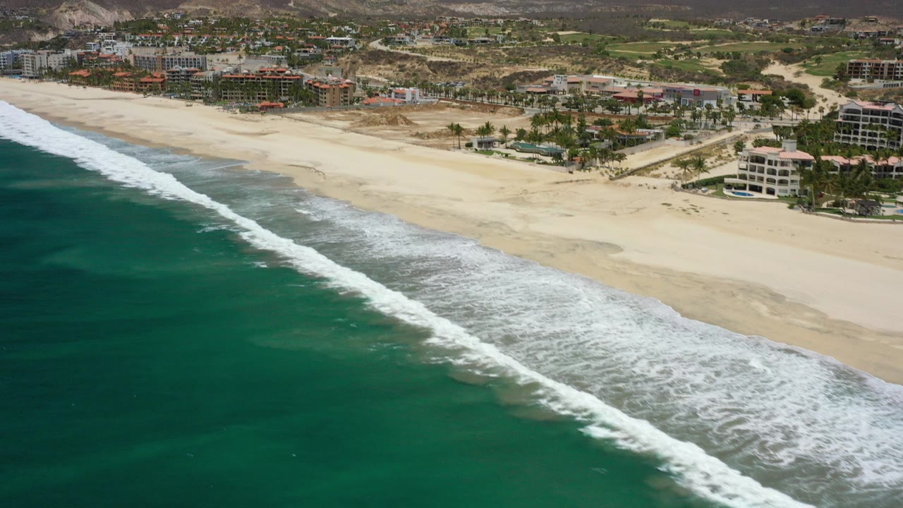 Aerial view of San Jose del Cabo beach with green-blue ocean in the foreground, a white surf band crossing the sand, beachfront buildings, dry hills, and no visible people.
