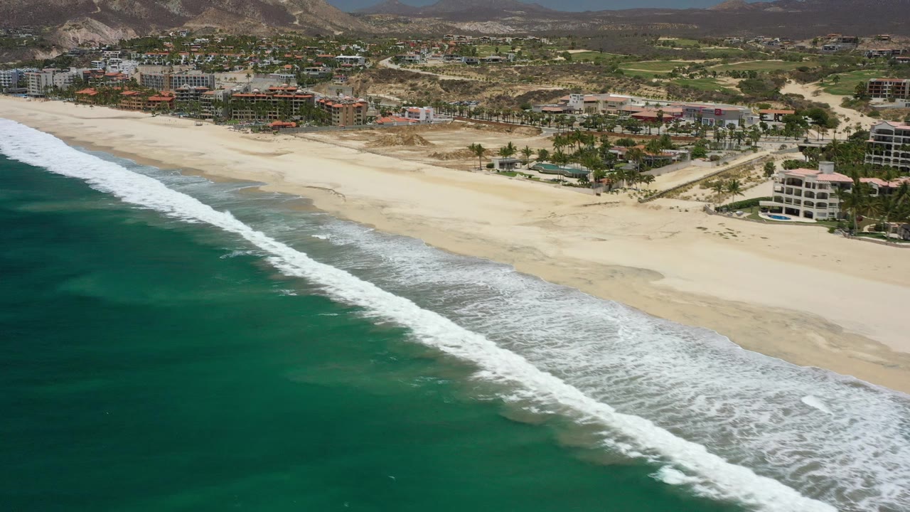 Wide aerial of beach, surf, shoreline buildings, green patches, and mountains beyond.