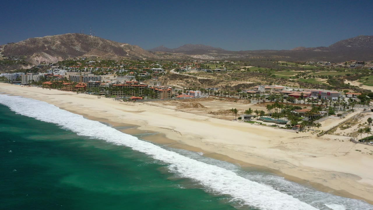 Aerial view of turquoise surf, wide sand beach, coastal buildings, and mountains.