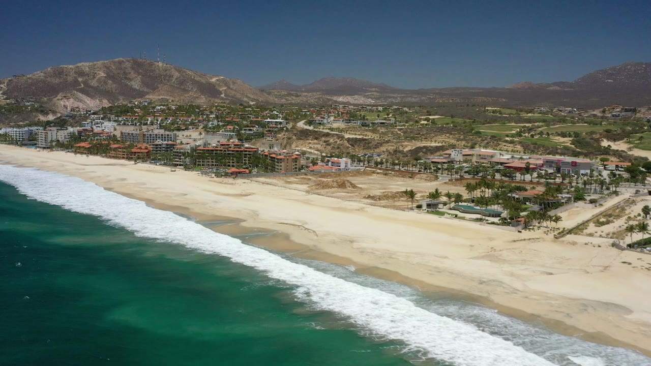 Aerial frame from San Jose del Cabo showing the surf band closer to the lower edge, open sand through the center, and a cluster of beachfront buildings backed by hills, with no visible people.