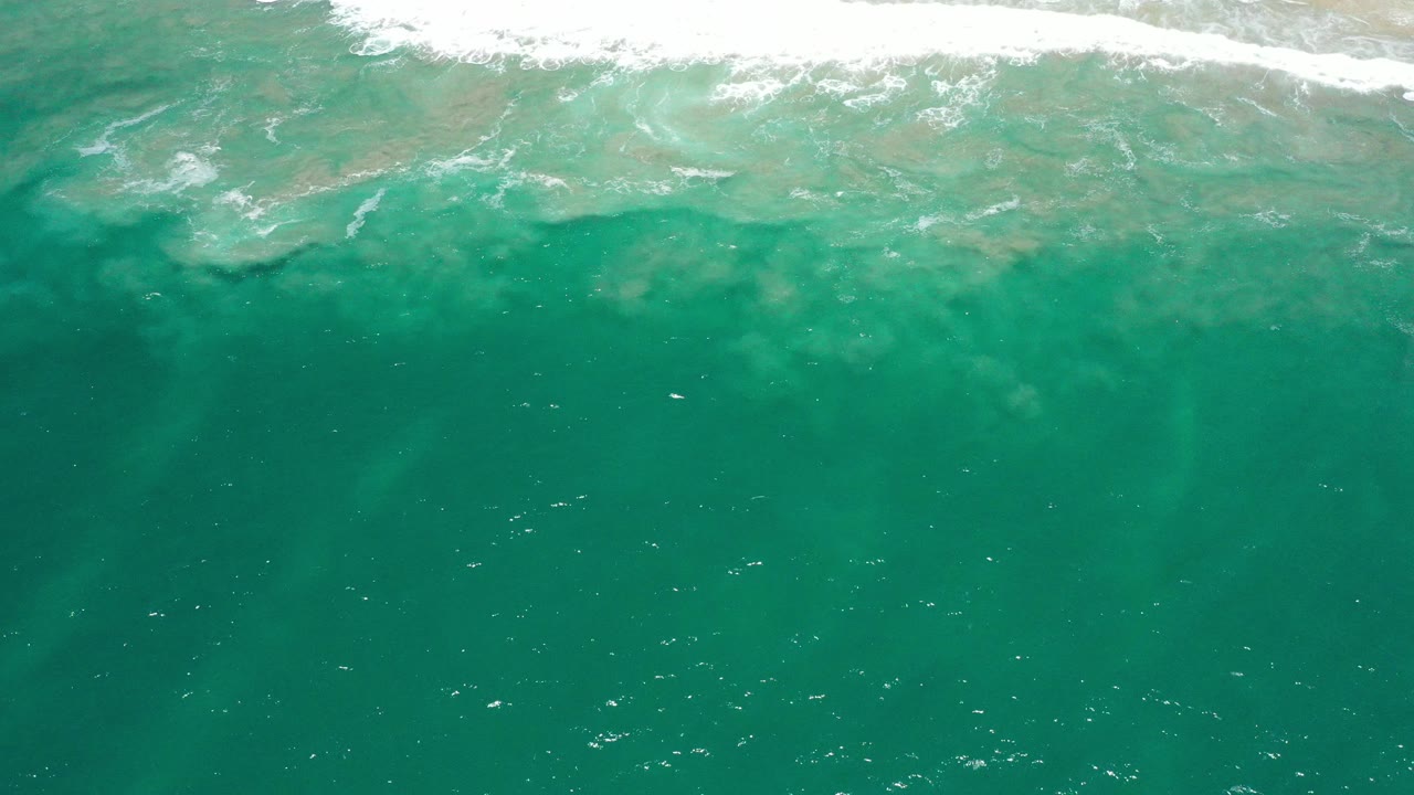 Overhead view of turquoise ocean water in San Jose Del Cabo, Mexico, with a white surf band near the top edge and no visible people.