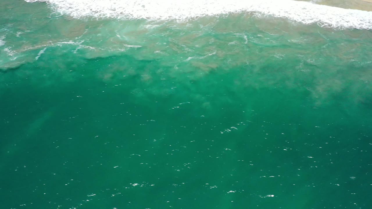 Aerial frame over deep green-turquoise water in San Jose del Cabo, with the foamy shore break running across the upper frame and no visible people.