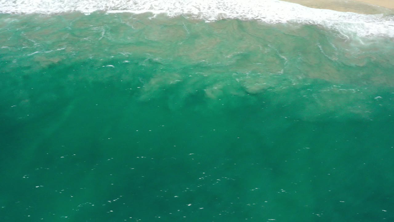 High aerial view of green-blue ocean water and a thin band of foamy surf meeting sand in San Jose Del Cabo, Mexico, with no visible people.