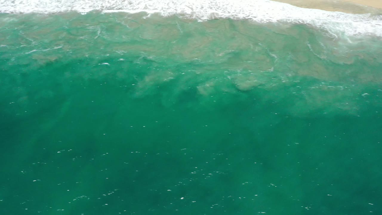 Aerial beach frame from San Jose Del Cabo, Mexico with turquoise water filling the foreground and white surf breaking near the sandy top edge, with no visible people.