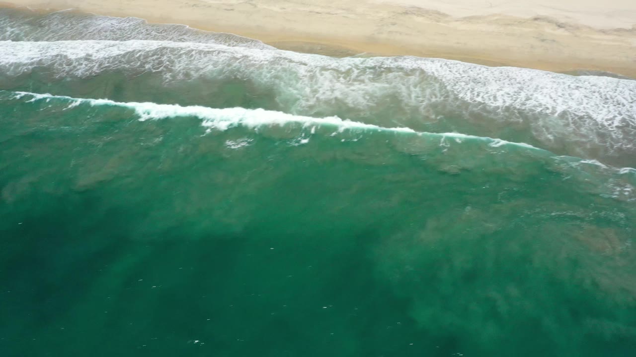 Aerial frame of white wave foam spreading across green ocean water beside the San Jose Del Cabo beach, with no visible people.