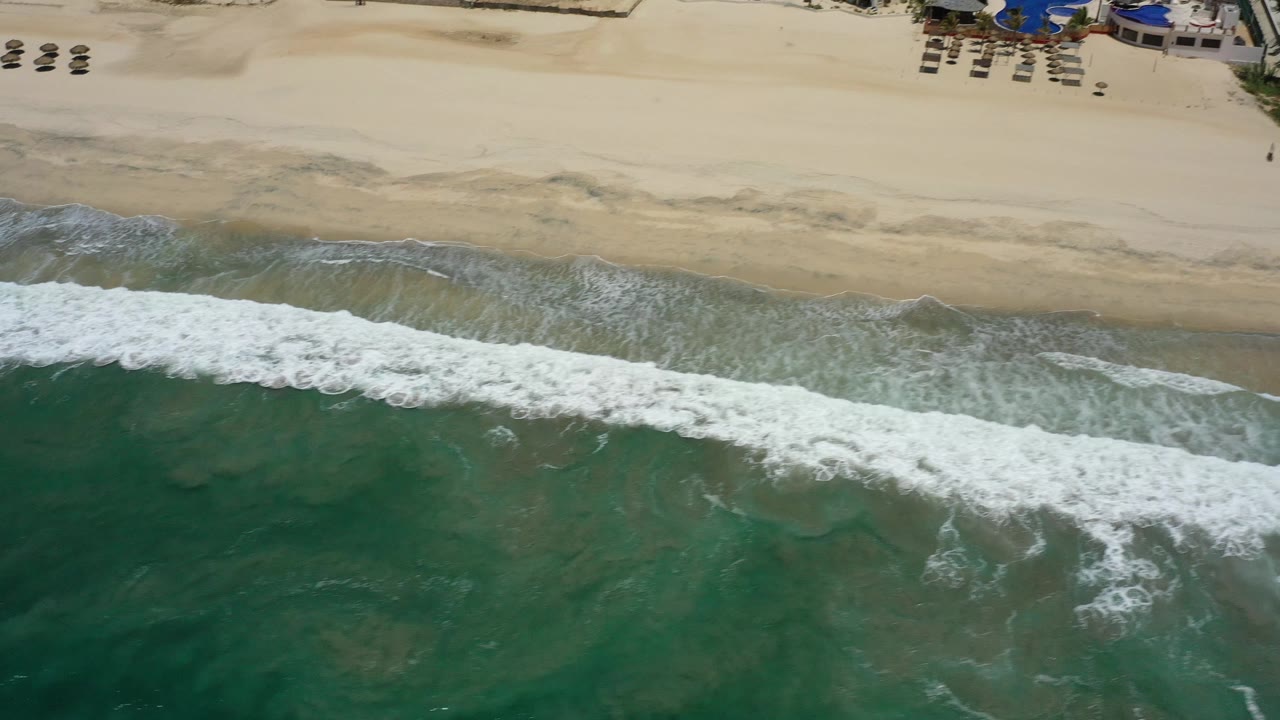 San Jose del Cabo beach from above, showing textured green water, foamy surf, broad tan sand, and small umbrella rows near the upper edge with no visible people.
