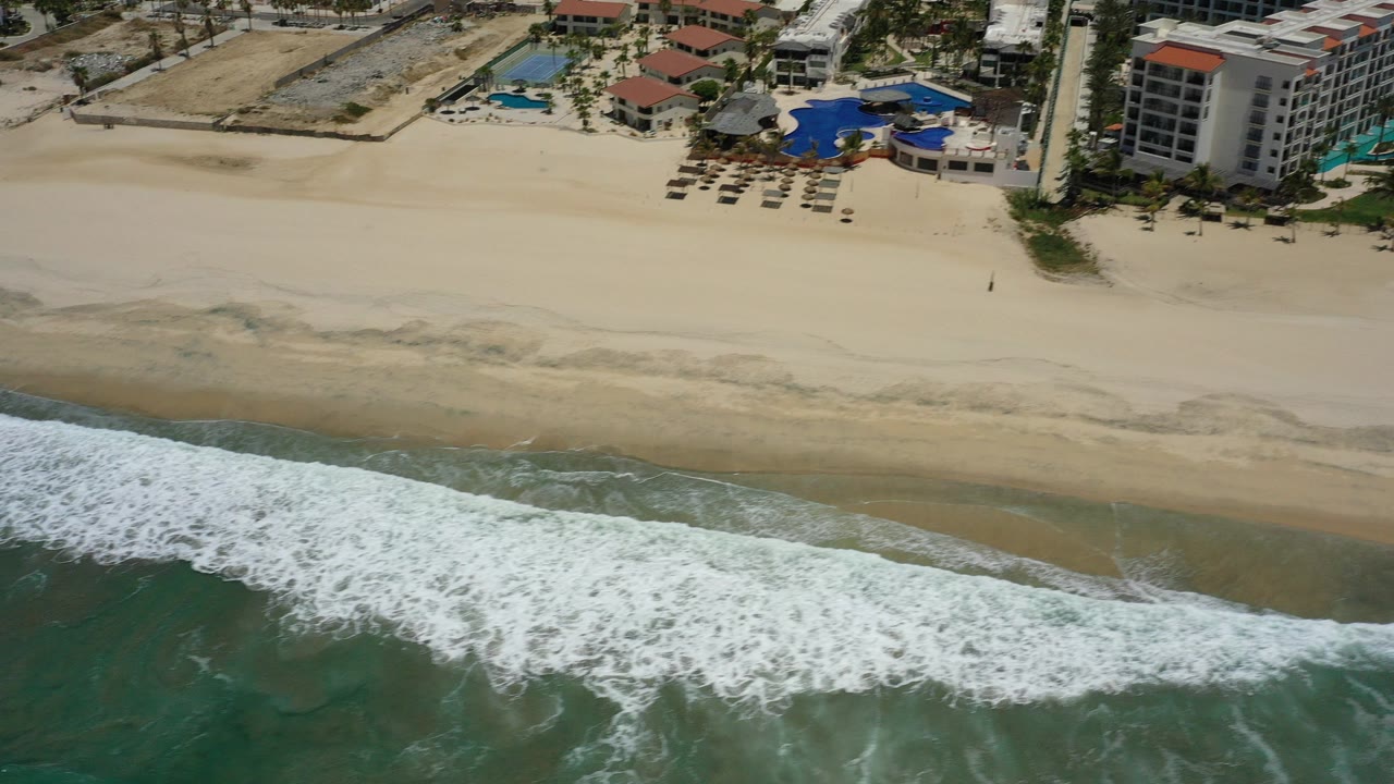 High aerial view in San Jose Del Cabo, Mexico, with green-blue surf in the foreground, a wide sand beach, resort pools and buildings at the top, and no visible people.