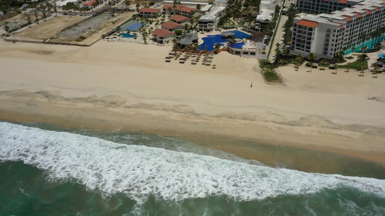 Overhead coastline with breaking waves and beachfront resort structures.
