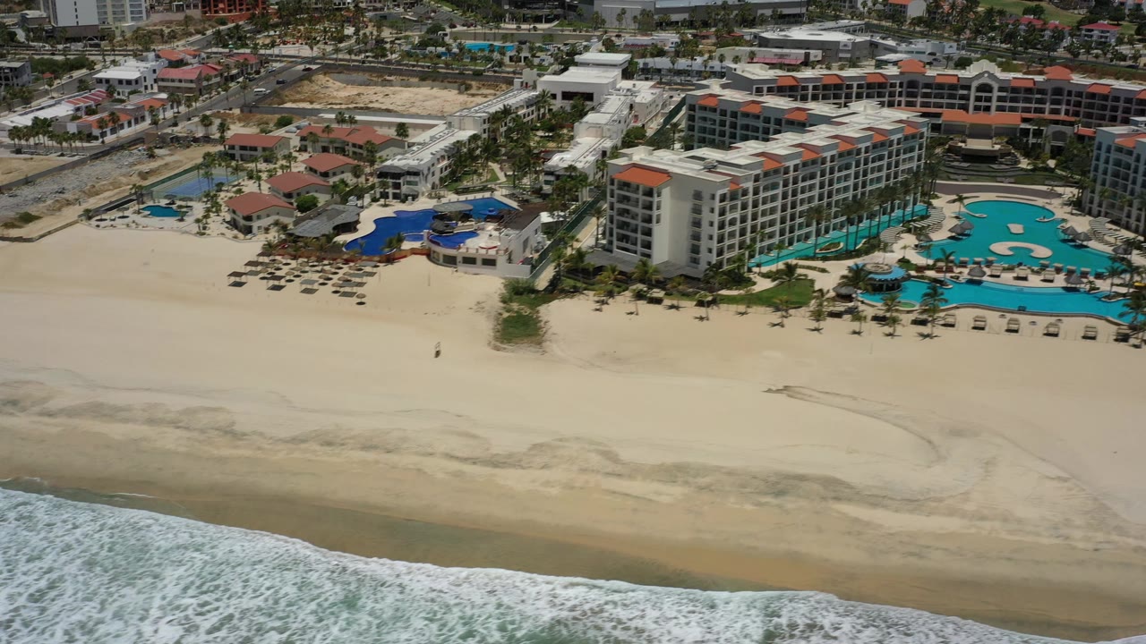Drone view in San Jose del Cabo, Mexico, showing a pale beach between foamy shoreline and resort pools, with hotel buildings and inland development behind, no visible people.