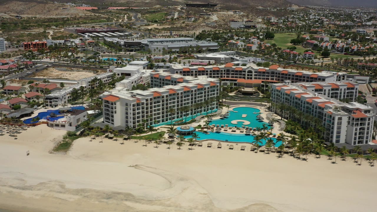 Aerial view of a beachfront resort with pools, palm trees, sand, town buildings, and hills.