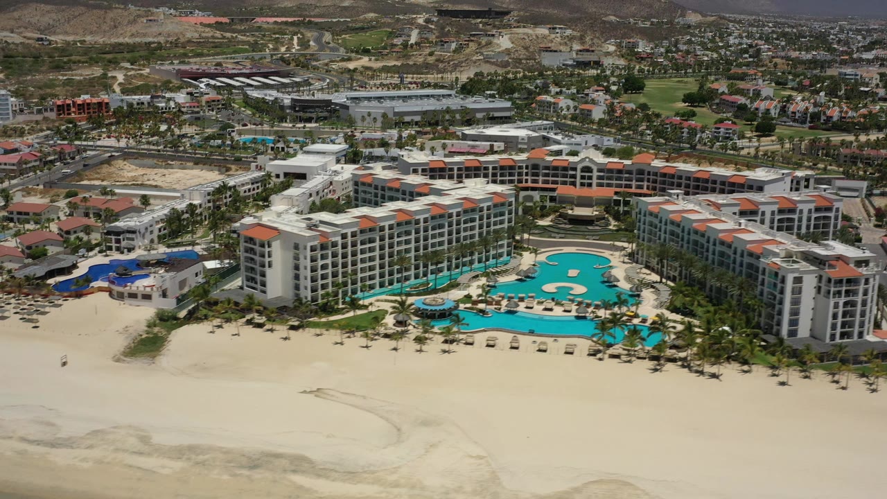 A second aerial frame in San Jose del Cabo shows the same beachfront resort shifted slightly right, with the central pool, rows of palms, adjacent buildings, arid hills, and no visible people.