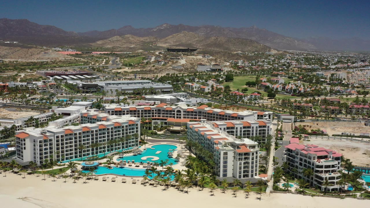 Aerial view of beachfront resort buildings, pools, sand, town, hills, and mountains.