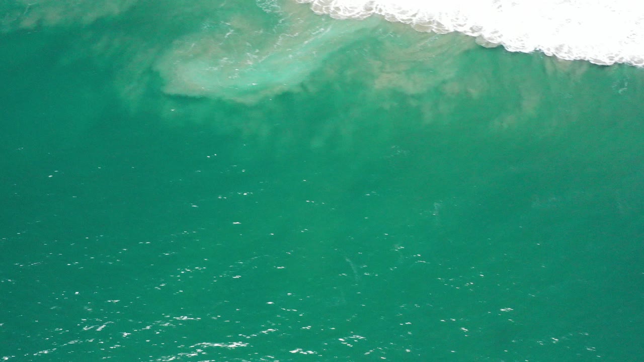 Aerial view of turquoise ocean in San Jose del Cabo, Mexico, with white surf at the top edge, pale submerged patches under the water, and no visible people.