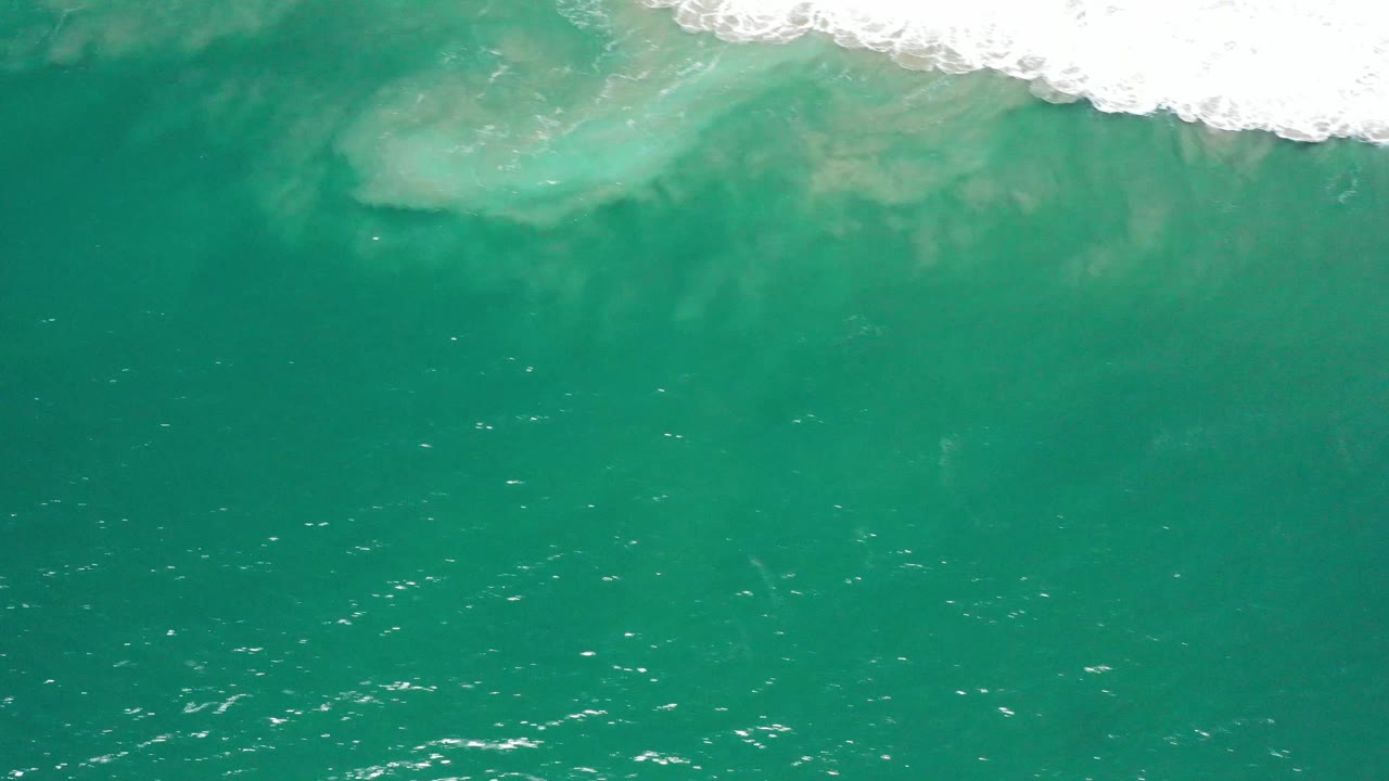 Aerial frame over blue-green water in San Jose del Cabo, Mexico, showing a pale submerged formation near the upper surf line and no visible people.