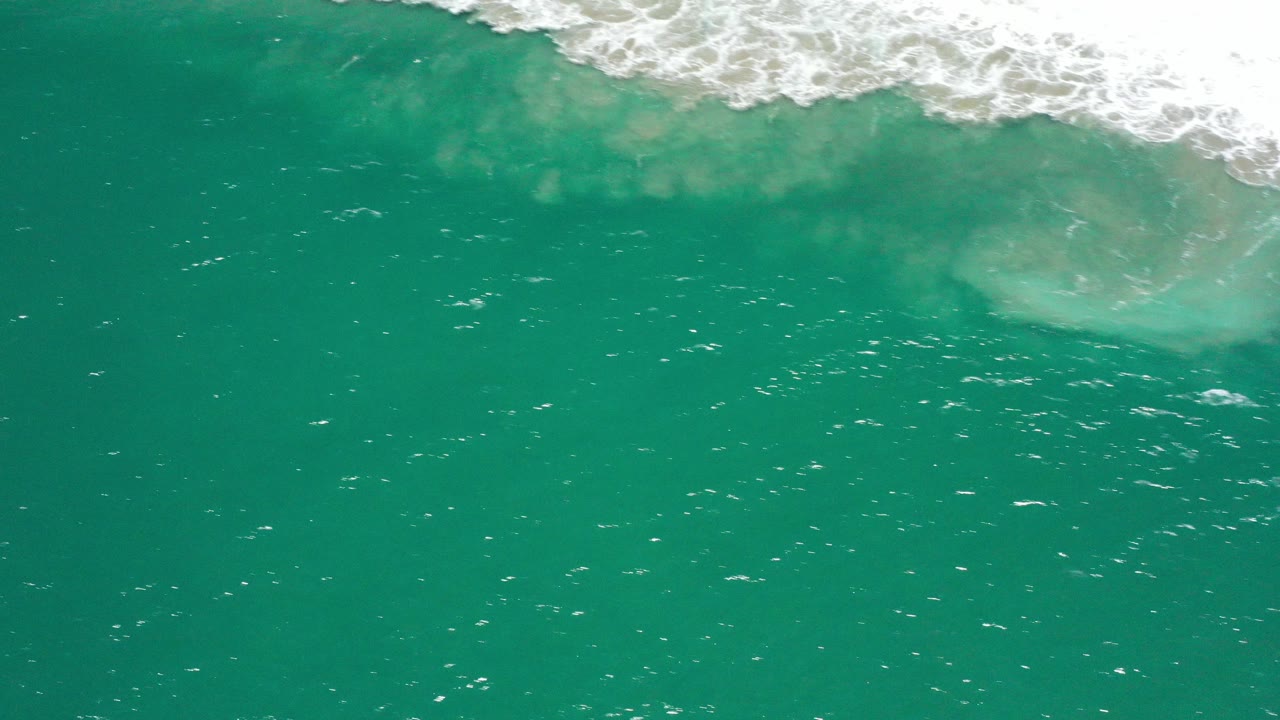 Overhead view of turquoise-green water in San Jose del Cabo with white surf foam entering along the top edge and no visible people.