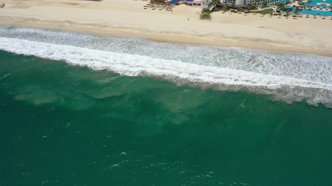 At 0:00, an aerial view in San Jose del Cabo shows deep green water in the foreground, a white surf band, pale sand, and distant beachfront buildings with no visible people.