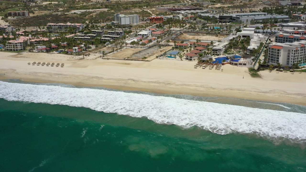 Aerial view in San Jose Del Cabo, Mexico, where a long band of white foam pushes toward the sand below beachfront hotels and arid hills, with no visible people.