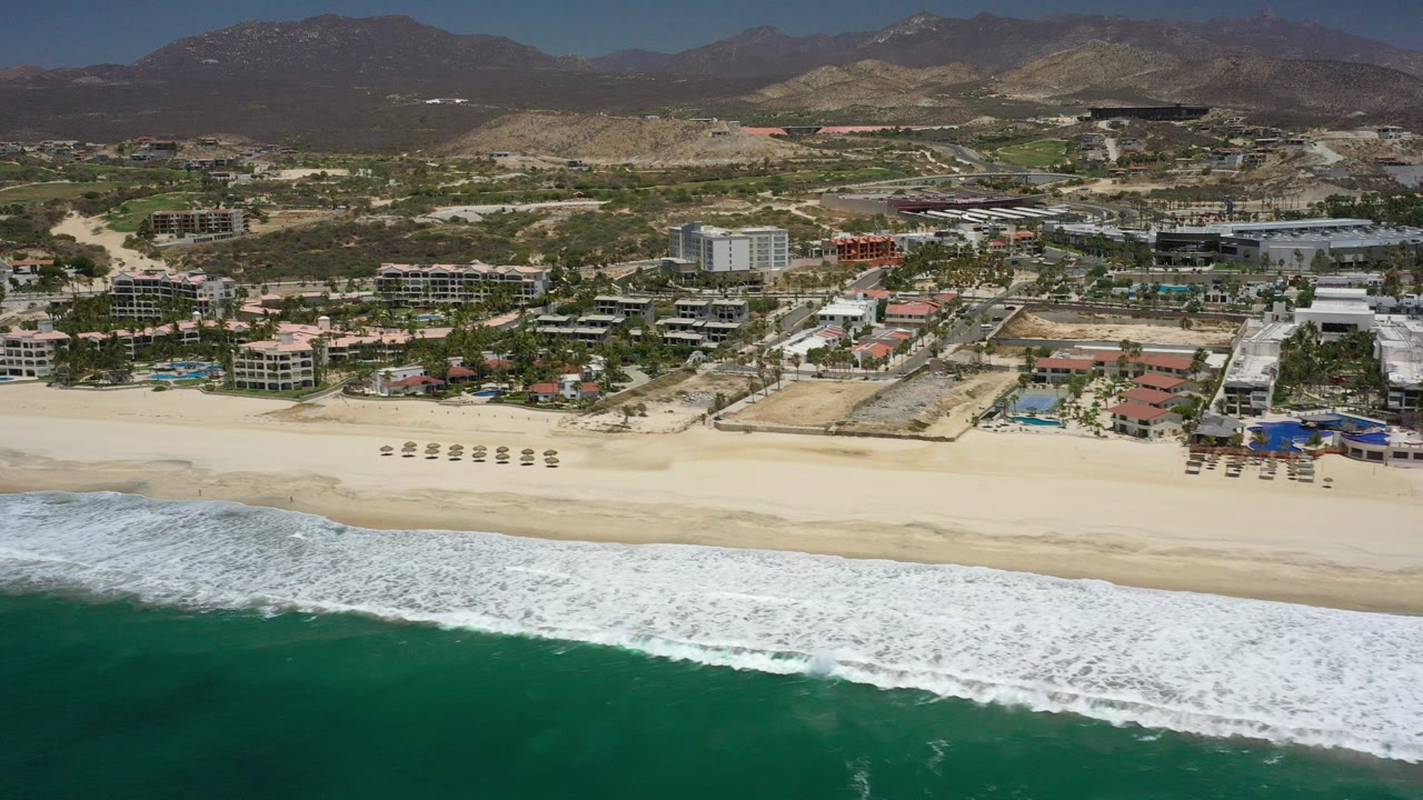 Aerial view of surf, sandy beach, resort buildings, and mountains.