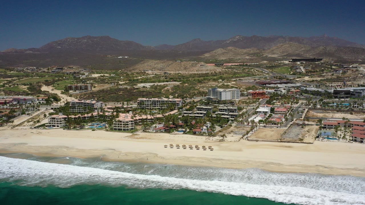 Wide aerial frame over San Jose del Cabo showing teal surf in the foreground, pale sand and beach umbrellas beyond it, resort buildings and roads inland, and no visible people.